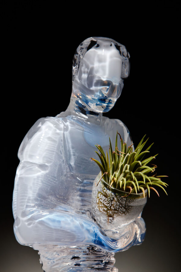 An opalescent glass figure carries a small metallic looking vessel which holds a small air plant. its features are blurred and unidentifiable but still convey sense of devotion and piety.