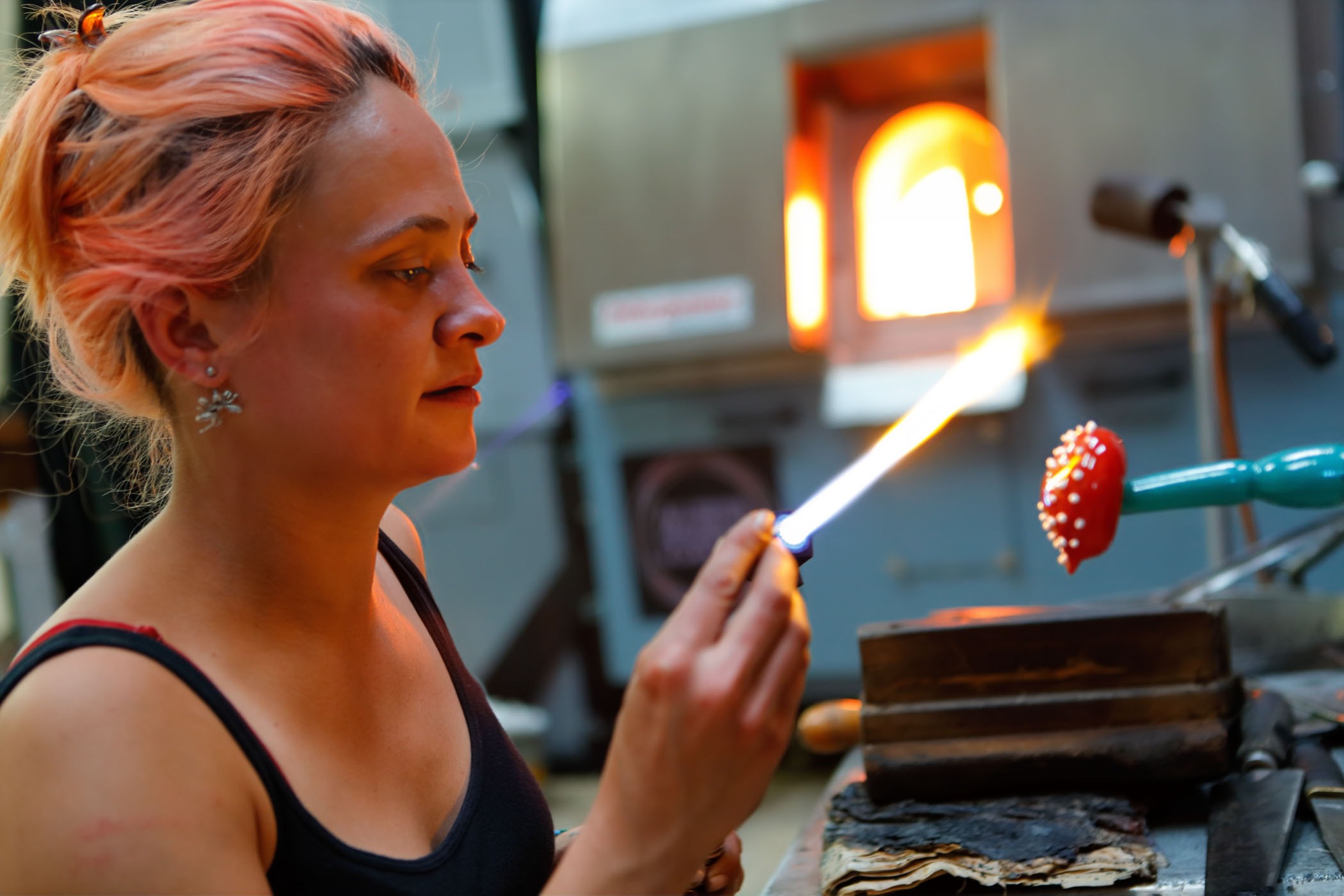 Madeleine hughes working in the glass studio at Stourbridge glass museum heats a small piece of glass to add detail to a small glass mushroom with the mouth of the glassfurnqace in the background